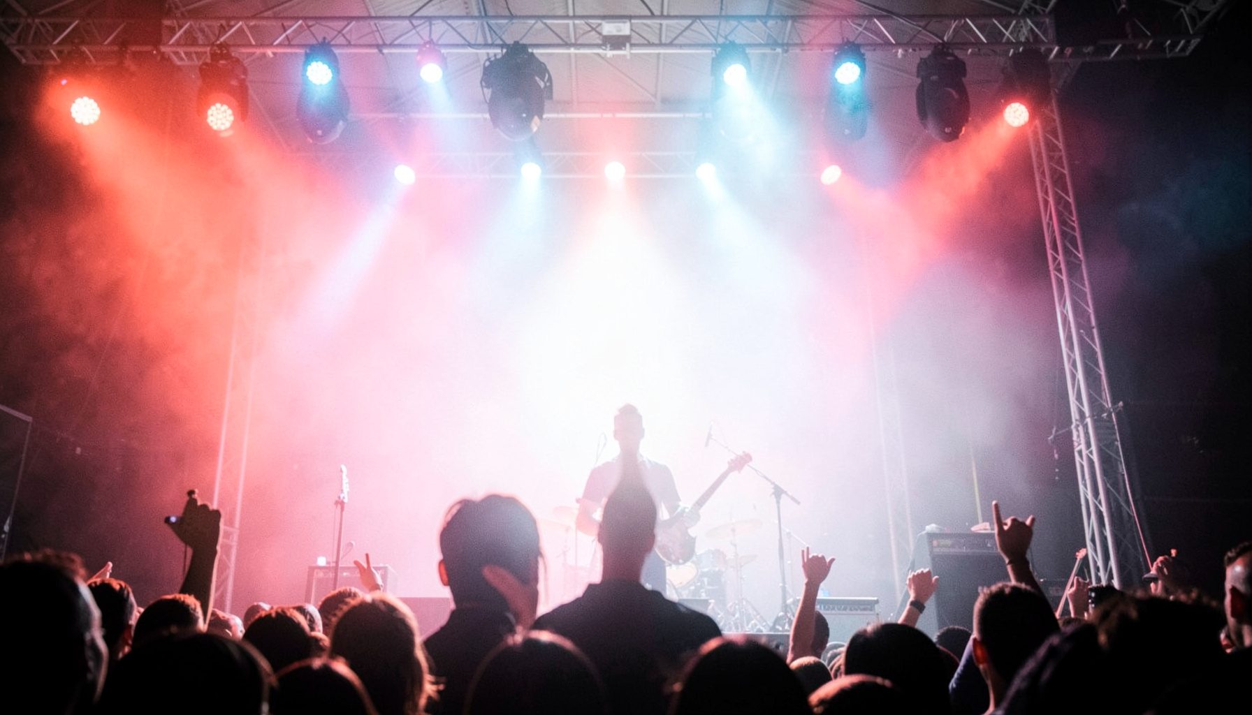 Palco de show com luzes vermelhas e azuis, silhueta de guitarra ao fundo, atmosfera intensa de rock ao vivo